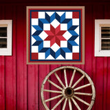 Carpenter Barn Quilt Pattern displayed on a rustic red barn with a vintage wagon wheel, showing the finished painted barn quilt outdoors.