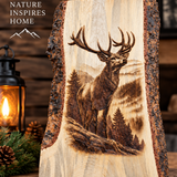 A rustic live-edge wooden sign featuring a detailed laser-burned or engraved stag in a mountain forest scene. The sign is displayed on a wooden surface next to a lit lantern and pinecones, with a wood-paneled background and the text 'NATURE INSPIRES HOME' in the upper left.