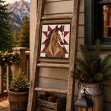 A rustic wooden ladder leans against a beige house exterior on a deck, displaying a geometric horse head art print. The scene is decorated with potted evergreens in galvanized buckets, pinecones in a wooden crate, and a lit lantern, with mountains and evergreens visible in the background.