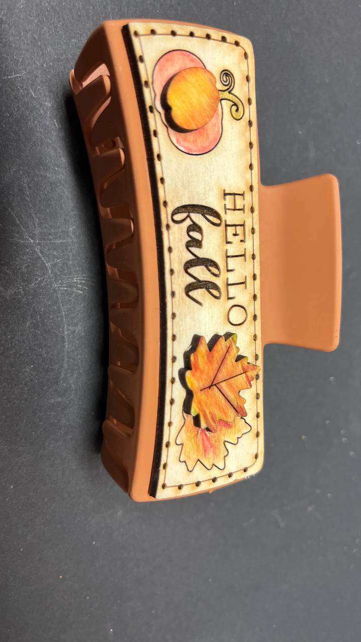A large wooden hair claw clip with a Hello Fall design, featuring an illustration of a pumpkin and fall leaves on the clip's clamp.