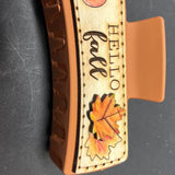 A large wooden hair claw clip with a Hello Fall design, featuring an illustration of a pumpkin and fall leaves on the clip's clamp.