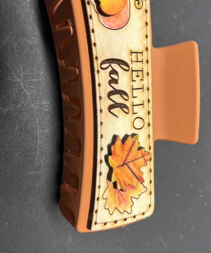 A large wooden hair claw clip with a Hello Fall design, featuring an illustration of a pumpkin and fall leaves on the clip's clamp.