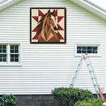 A square framed wall art print featuring a stylized brown horse head on a cream and burgundy geometric background, mounted on the exterior of a white house above two windows, with a ladder leaning nearby.