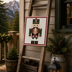 A framed nutcracker needlepoint canvas displayed on a rustic wooden ladder on a porch. The scene is decorated with potted evergreens in a galvanized bucket, pinecones in a wooden crate, a lit lantern, and mountains in the background.