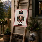 A framed nutcracker needlepoint canvas displayed on a rustic wooden ladder on a porch. The scene is decorated with potted evergreens in a galvanized bucket, pinecones in a wooden crate, a lit lantern, and mountains in the background.