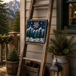 A rustic wooden ladder leans against a house exterior on a porch, displaying a square decorative tile with a winter mountain scene. The scene includes pine branches in a galvanized bucket, a black lantern with a lit candle, and mountains in the background.