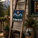 A rustic wooden ladder leans against a house exterior on a porch, displaying a square decorative tile with a winter mountain scene. The scene includes pine branches in a galvanized bucket, a black lantern with a lit candle, and mountains in the background.
