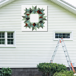 A square wall art print featuring a geometric starburst pattern in muted green, gray, black, and brown tones, displayed on the exterior of a white-sided house above a window, with a ladder nearby.
