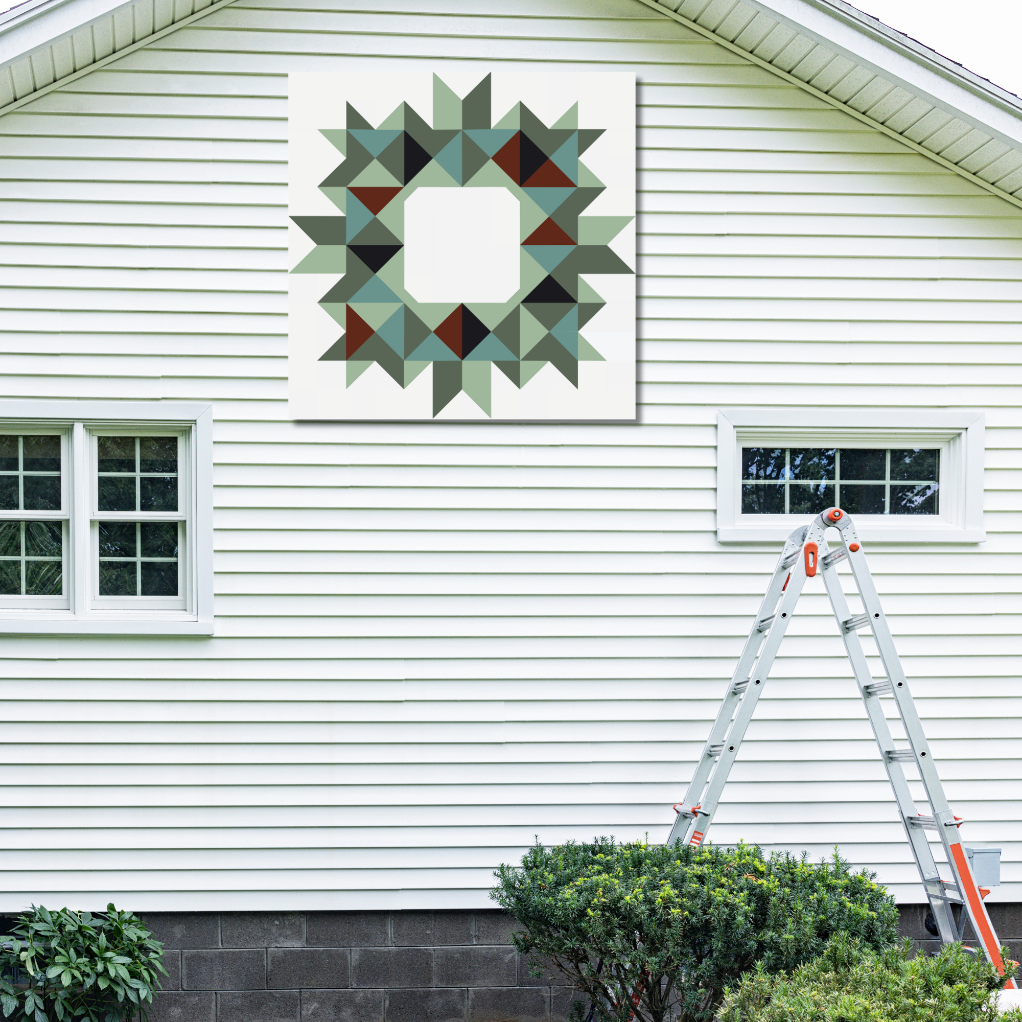 A square wall art print featuring a geometric starburst pattern in muted green, gray, black, and brown tones, displayed on the exterior of a white-sided house above a window, with a ladder nearby.