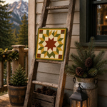 A rustic wooden ladder leans against a house exterior on a porch, displaying a square quilted tile with a geometric star pattern in green, yellow, cream, and brown. The scene includes potted evergreens in galvanized buckets, pinecones in a wooden crate, a lit lantern, and mountains in the background.