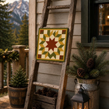 A rustic wooden ladder leans against a house exterior on a porch, displaying a square quilted tile with a geometric star pattern in green, yellow, cream, and brown. The scene includes potted evergreens in galvanized buckets, pinecones in a wooden crate, a lit lantern, and mountains in the background.