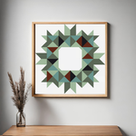 Framed geometric wall art hanging on a light gray wall above a wooden console table, with a glass vase of dried grass on the left. The artwork features a circular starburst pattern made of many triangles in muted green, teal, dark green, brown, and black on a white background, enclosed in a light natural wood frame.