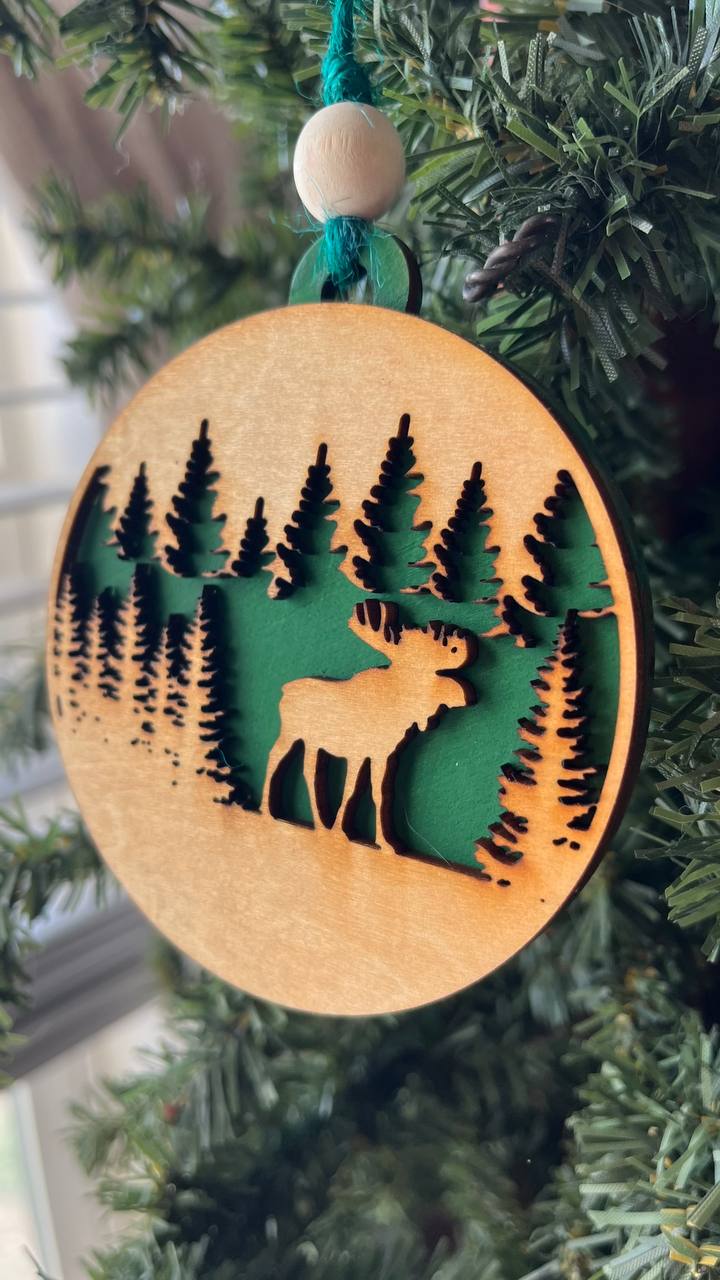 A round wooden Christmas ornament featuring a laser-cut moose and pine tree design, hanging from a green string on a Christmas tree branch.