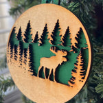 A round wooden Christmas ornament featuring a laser-cut moose and pine tree design, hanging from a green string on a Christmas tree branch.
