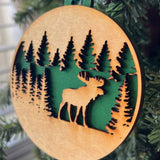 A round wooden Christmas ornament featuring a laser-cut moose and pine tree design, hanging from a green string on a Christmas tree branch.
