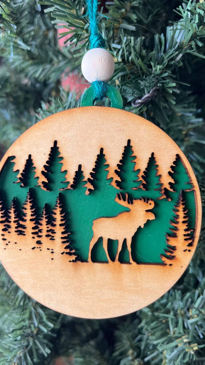 A handcrafted wooden ornament featuring a moose among trees, with a circular design and natural wood finish. The ornament includes a stained top layer for detail and a hand-painted back layer, available in various colors like red, orange, yellow, green, blue, or an ombre effect. It measures 3.25 inches in diameter and comes with a twine hanger and natural color bead, offering optional engraving for personalization with a name or date. Ideal for rustic holiday decor and personalized gifts.

