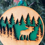 A handcrafted wooden ornament featuring a moose among trees, with a circular design and natural wood finish. The ornament includes a stained top layer for detail and a hand-painted back layer, available in various colors like red, orange, yellow, green, blue, or an ombre effect. It measures 3.25 inches in diameter and comes with a twine hanger and natural color bead, offering optional engraving for personalization with a name or date. Ideal for rustic holiday decor and personalized gifts.

