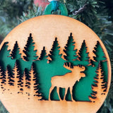 A handcrafted wooden ornament featuring a moose among trees, with a circular design and natural wood finish. The ornament includes a stained top layer for detail and a hand-painted back layer, available in various colors like red, orange, yellow, green, blue, or an ombre effect. It measures 3.25 inches in diameter and comes with a twine hanger and natural color bead, offering optional engraving for personalization with a name or date. Ideal for rustic holiday decor and personalized gifts.

