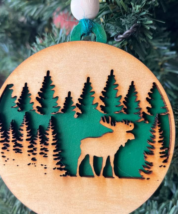 A handcrafted wooden ornament featuring a moose among trees, with a circular design and natural wood finish. The ornament includes a stained top layer for detail and a hand-painted back layer, available in various colors like red, orange, yellow, green, blue, or an ombre effect. It measures 3.25 inches in diameter and comes with a twine hanger and natural color bead, offering optional engraving for personalization with a name or date. Ideal for rustic holiday decor and personalized gifts.

