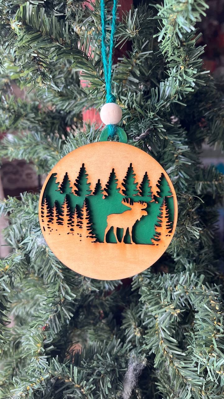 A handcrafted wooden ornament featuring a moose among trees, with a circular design and natural wood finish. The ornament includes a stained top layer for detail and a hand-painted back layer, available in various colors like red, orange, yellow, green, blue, or an ombre effect. It measures 3.25 inches in diameter and comes with a twine hanger and natural color bead, offering optional engraving for personalization with a name or date. Ideal for rustic holiday decor and personalized gifts.

