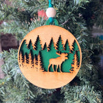 A handcrafted wooden ornament featuring a moose among trees, with a circular design and natural wood finish. The ornament includes a stained top layer for detail and a hand-painted back layer, available in various colors like red, orange, yellow, green, blue, or an ombre effect. It measures 3.25 inches in diameter and comes with a twine hanger and natural color bead, offering optional engraving for personalization with a name or date. Ideal for rustic holiday decor and personalized gifts.

