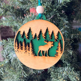 A handcrafted wooden ornament featuring a moose among trees, with a circular design and natural wood finish. The ornament includes a stained top layer for detail and a hand-painted back layer, available in various colors like red, orange, yellow, green, blue, or an ombre effect. It measures 3.25 inches in diameter and comes with a twine hanger and natural color bead, offering optional engraving for personalization with a name or date. Ideal for rustic holiday decor and personalized gifts.


