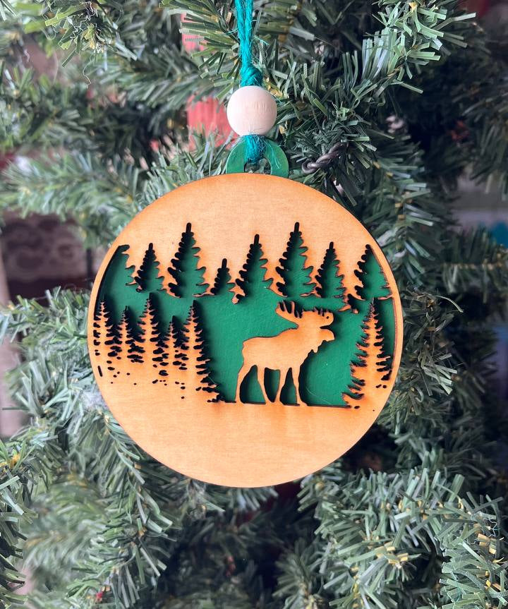 A handcrafted wooden ornament featuring a moose among trees, with a circular design and natural wood finish. The ornament includes a stained top layer for detail and a hand-painted back layer, available in various colors like red, orange, yellow, green, blue, or an ombre effect. It measures 3.25 inches in diameter and comes with a twine hanger and natural color bead, offering optional engraving for personalization with a name or date. Ideal for rustic holiday decor and personalized gifts.

