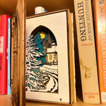 Handmade wooden Christmas book nook featuring a layered winter church, snow-covered evergreen trees and moonlit landscape for rustic Christmas bookshelf decor.