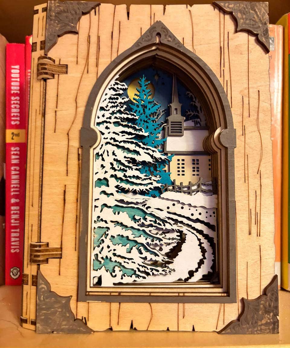 Handmade wooden Christmas book nook featuring a layered winter church, snow-covered evergreen trees and moonlit landscape for rustic Christmas bookshelf decor.