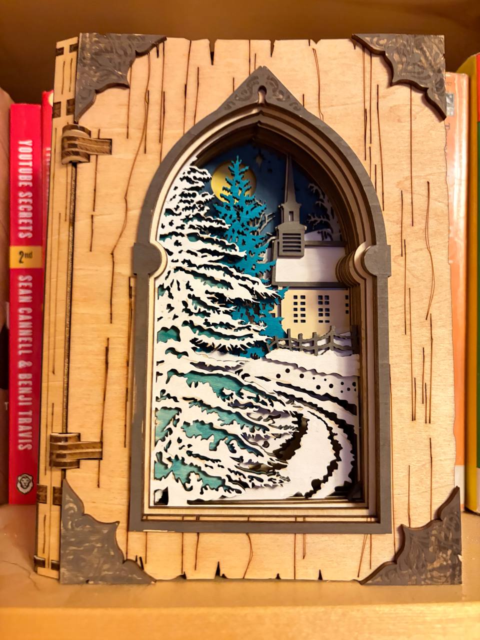 Handmade wooden Christmas book nook featuring a layered winter church, snow-covered evergreen trees and moonlit landscape for rustic Christmas bookshelf decor.