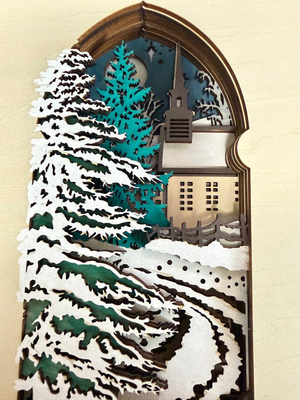 Handmade wooden Christmas book nook featuring a layered winter church, snow-covered evergreen trees and moonlit landscape for rustic Christmas bookshelf decor.