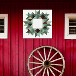 A square wall art print featuring a geometric circular pattern in shades of green, blue, and brown on a white background, displayed on a red wooden barn wall above a large rustic wheel.